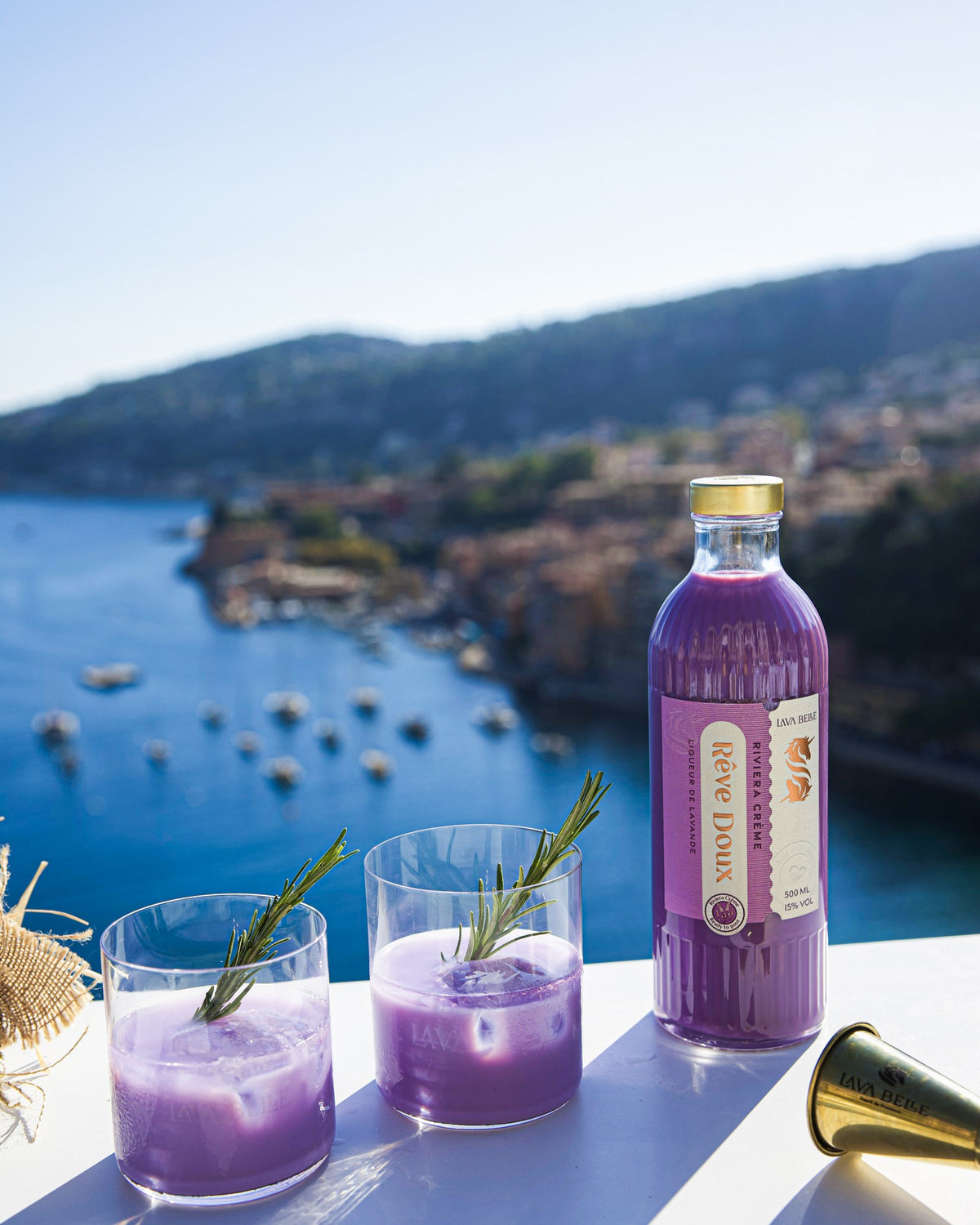Fles Lav'a Belle Rêve Doux en twee glazen met een romig paars drankje en rozemarijn, op de achtergrond een baai aan de Côte d'Azur