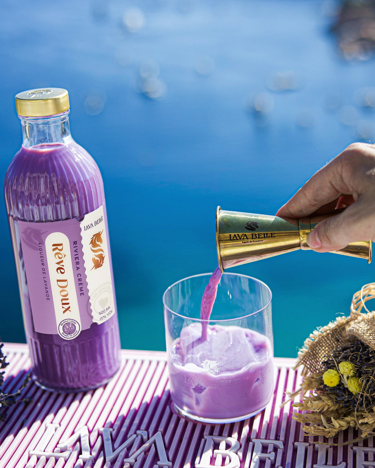 Romige paarse Rêve Doux wordt uit de jigger in het glas geschonken, ernaast de fles en een boeket droogbloemen